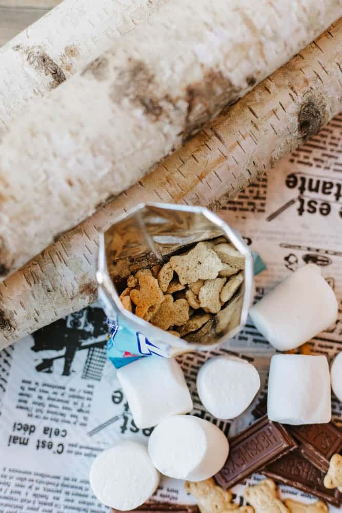 An opened bag of bear-shaped graham crackers, marshmallows, and chocolate pieces on newspaper, with two birch logs placed nearby.