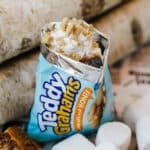 An opened bag of Teddy Grahams filled with whipped cream, crushed cookies, and chocolate, surrounded by marshmallows and toasted marshmallows on a stick, with birch logs in the background.