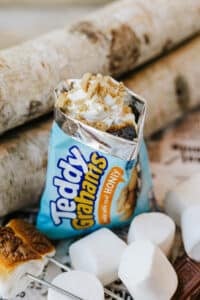 An opened bag of Teddy Grahams filled with whipped cream, crushed cookies, and chocolate, surrounded by marshmallows and toasted marshmallows on a stick, with birch logs in the background.