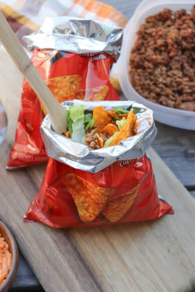 An open bag of Doritos with taco toppings, including lettuce, cheese, and ground beef, being mixed with a wooden spoon. Another bag and a container of seasoned ground beef are in the background on a wooden surface.