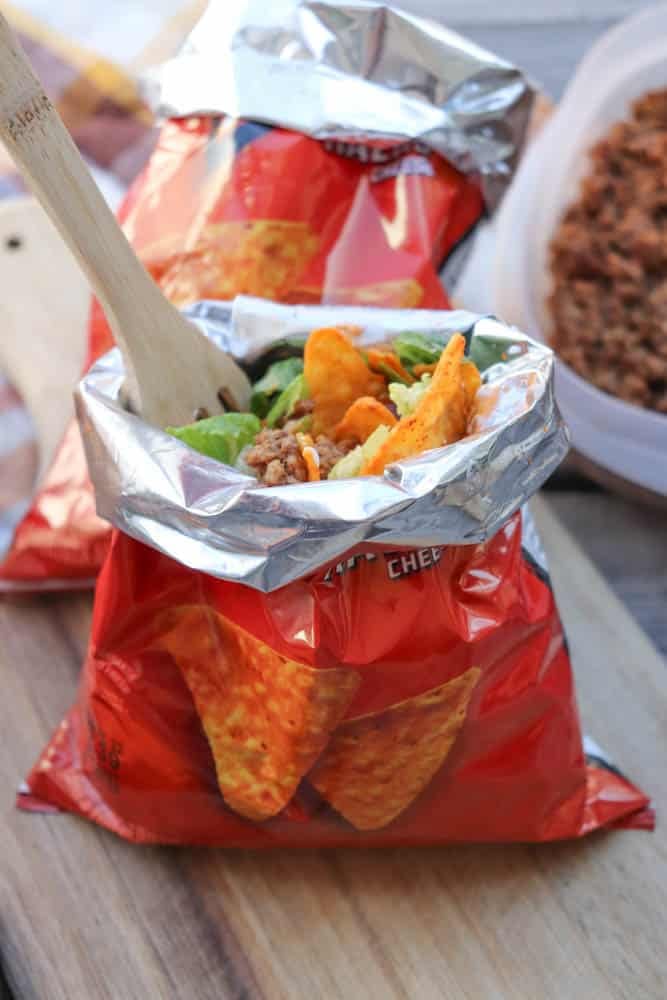 An open bag of Doritos used as a container, filled with taco ingredients like ground meat, lettuce, and cheese, with a wooden fork inside. A tray of more taco filling is in the background.