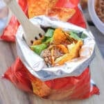 An open bag of nacho cheese chips filled with taco ingredients like ground beef, lettuce, cheese, and salsa, with a wooden fork inside, sitting on a wooden surface.