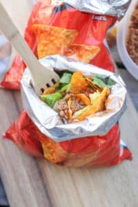 An open bag of nacho cheese chips filled with taco ingredients like ground beef, lettuce, cheese, and salsa, with a wooden fork inside, sitting on a wooden surface.