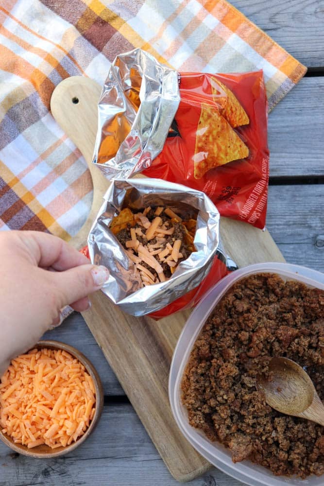 A hand holds an open snack-size Doritos bag filled with shredded cheese above a wooden board, next to taco meat, a bowl of cheese, and a plaid cloth on a wooden table.