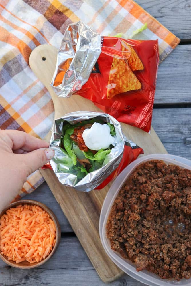 A hand holds an open Doritos chip bag filled with taco ingredients like lettuce and sour cream. Nearby are ground beef, shredded cheese, and another bag of Doritos on a wooden board and plaid towel.