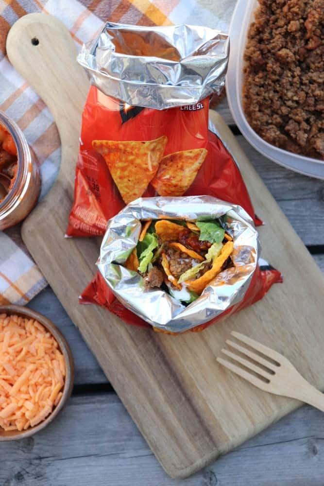 A small bag of Doritos opened and filled with taco meat, lettuce, cheese, and salsa, sits on a wooden board next to a bowl of shredded cheese and a container of seasoned ground beef.