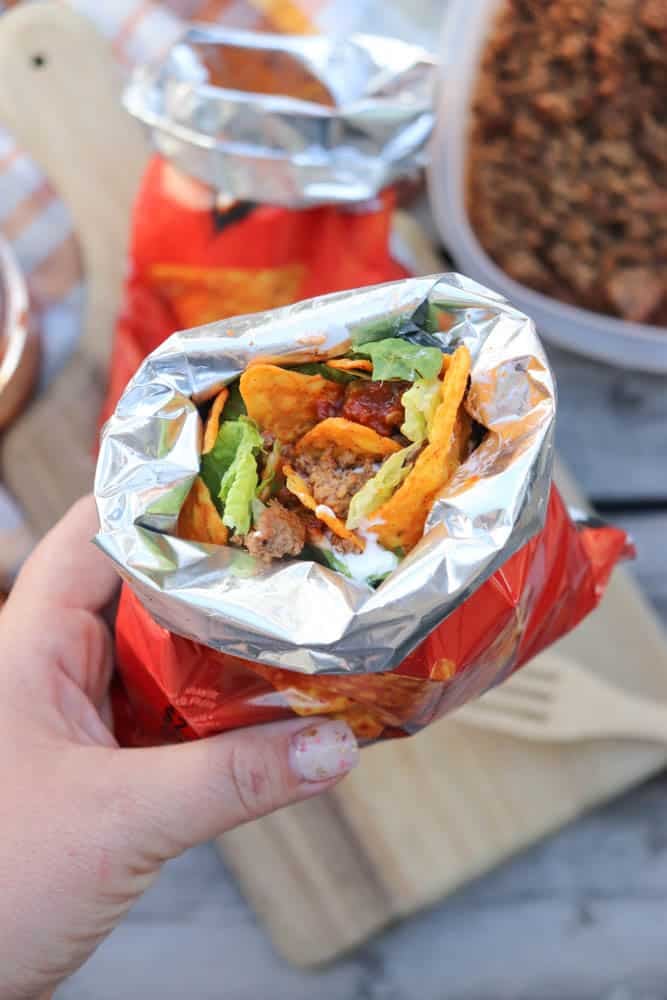 A hand holds an open chip bag filled with taco ingredients like ground beef, lettuce, cheese, and chips, creating a portable taco salad. A fork, cutting board, and more food are visible in the background.