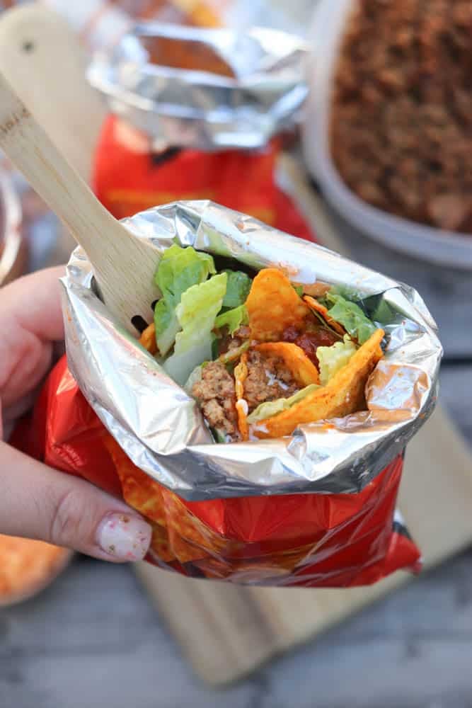 A hand holds an open snack-size chip bag filled with taco ingredients like lettuce, ground beef, cheese, and chips, using a wooden fork.