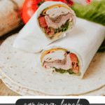 A close-up of two halved deli meat and veggie wraps on a tortilla, with lettuce, tomato, and cheese visible, placed on a wooden surface. Text below reads: Camping Lunch Wraps Recipe campfirefoodie.com.