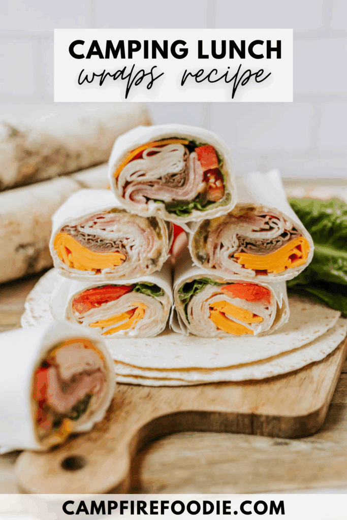 A stack of colorful sandwich wraps filled with deli meat, cheese, lettuce, and tomato are displayed on a rustic wooden board, with text reading Camping Lunch Wraps Recipe and campfirefoodie.com.