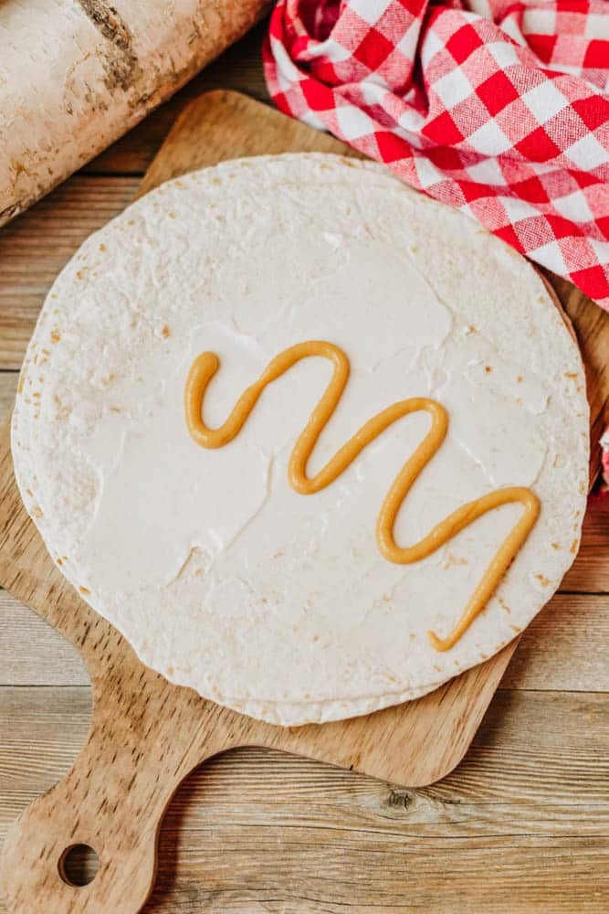 A flour tortilla spread with cream cheese and topped with a zigzag line of honey mustard sits on a wooden cutting board next to a red and white checkered cloth.