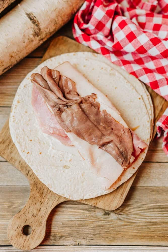 A tortilla on a wooden board topped with slices of roast beef and ham, next to a red and white checkered cloth and pieces of birch wood on a rustic wooden surface.