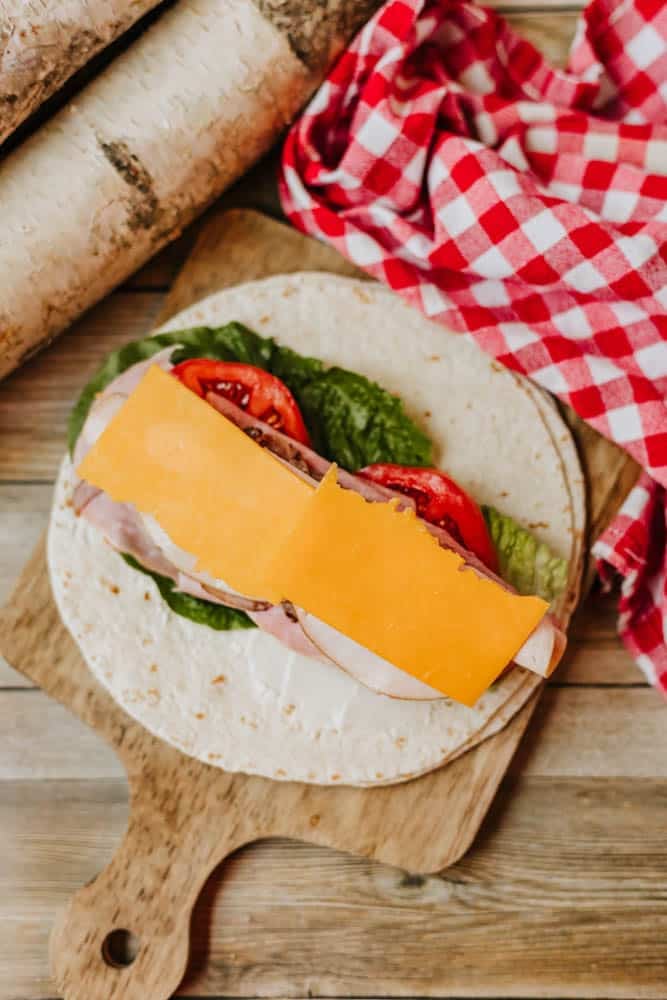 A tortilla wrap on a wooden board, layered with lettuce, tomato slices, sliced turkey, and a slice of cheddar cheese, next to a red and white checkered cloth and birch logs.