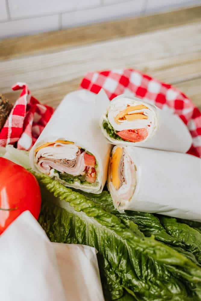 Three tortilla wraps filled with deli meat, cheddar cheese, lettuce, and tomato, wrapped in white paper, are displayed on romaine lettuce next to a red-and-white checkered cloth and a fresh tomato.