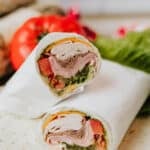 A close-up of a sandwich wrap cut in half, showing layers of sliced turkey, cheese, lettuce, and tomato wrapped in a flour tortilla, with fresh vegetables in the background.