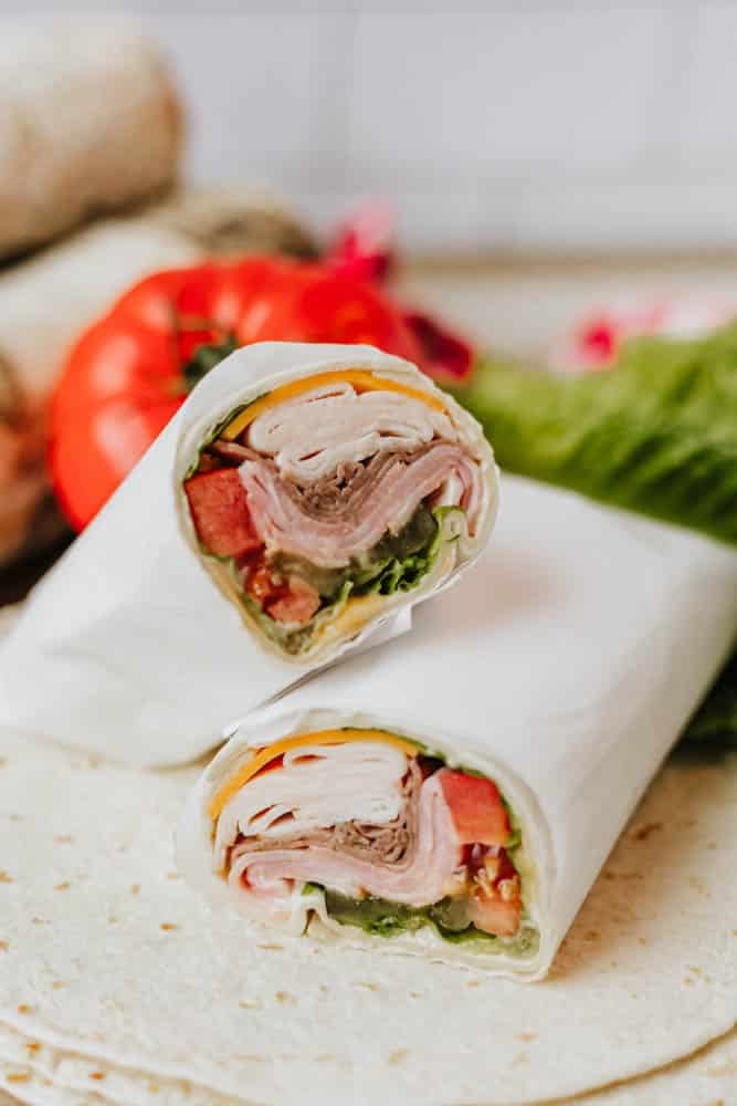 A close-up of a sandwich wrap cut in half, showing layers of sliced turkey, cheese, lettuce, and tomato wrapped in a flour tortilla, with fresh vegetables in the background.