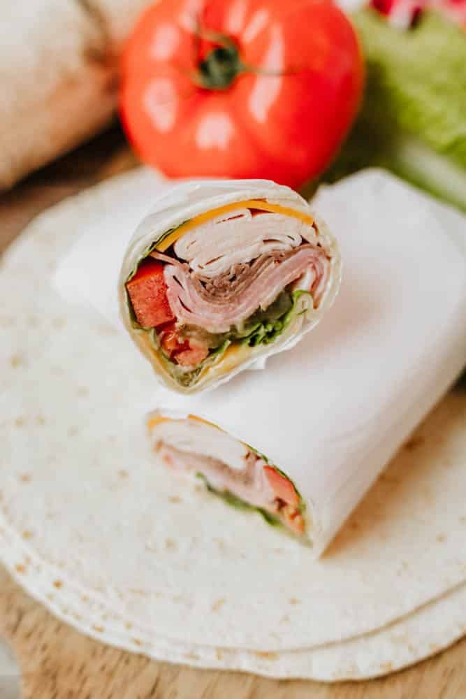 A sliced turkey and vegetable wrap on a tortilla, filled with lettuce, tomato, and deli meat, wrapped in white paper. A fresh tomato and lettuce are blurred in the background.