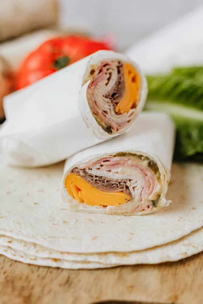 A close-up of a tortilla wrap cut in half, filled with sliced deli meats, cheddar cheese, and pickles, with lettuce, a tomato, and tortilla wraps in the background.