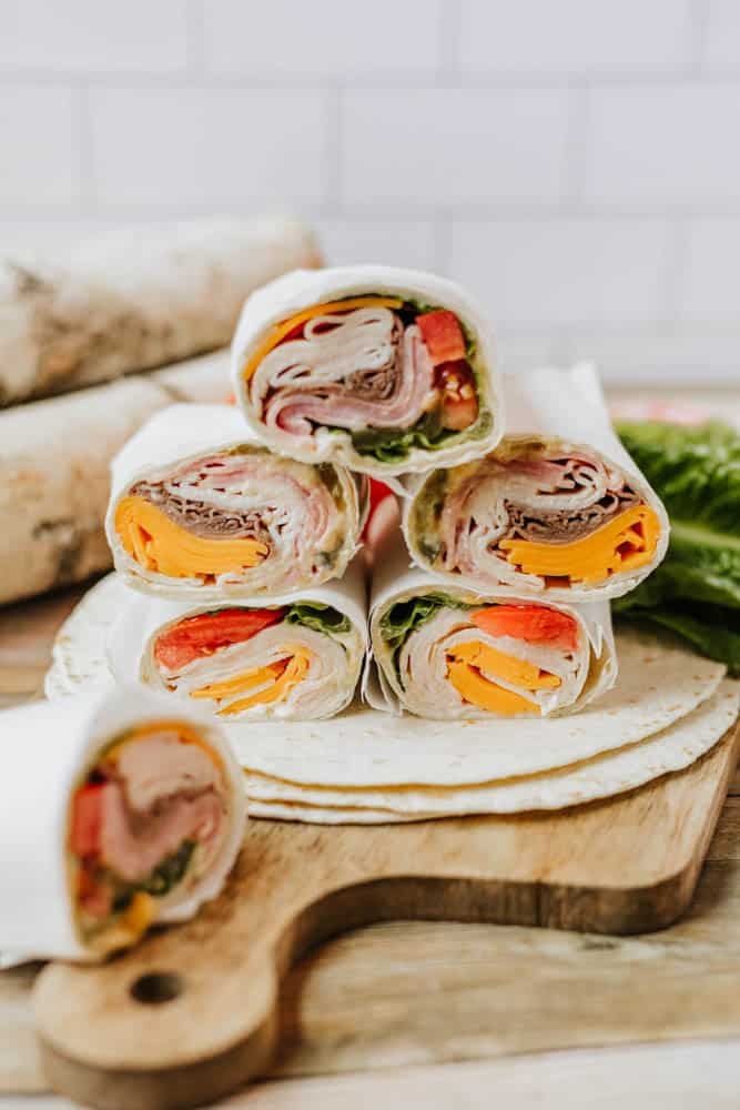 Six colorful deli meat wraps filled with lettuce, cheese, and tomatoes are stacked on a wooden board. The wraps are sliced to show the layers of ingredients, and a few tortillas and lettuce are in the background.