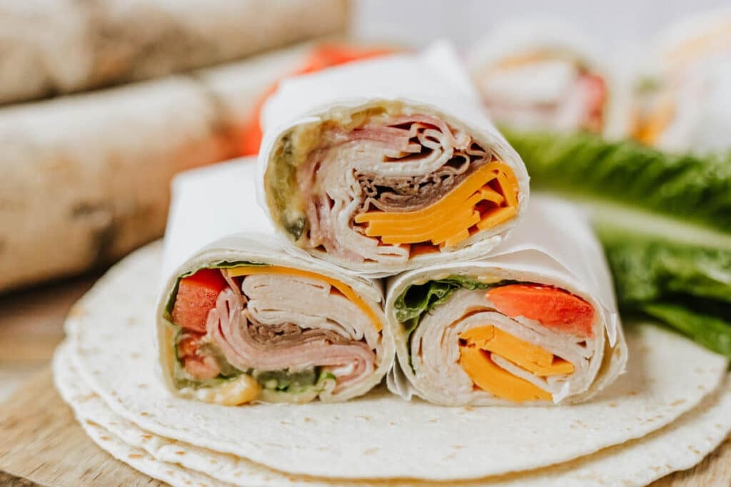 Three tortilla wraps filled with sliced deli meats, cheddar cheese, lettuce, and tomato are stacked on top of each other, with a fresh lettuce leaf and tortillas in the background.