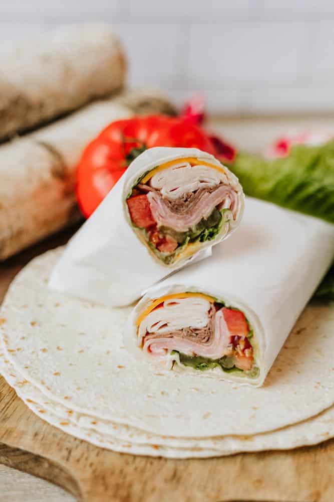 A tortilla wrap cut in half, filled with deli meat, cheese, lettuce, and tomato, sits on a wooden board with fresh vegetables in the background.