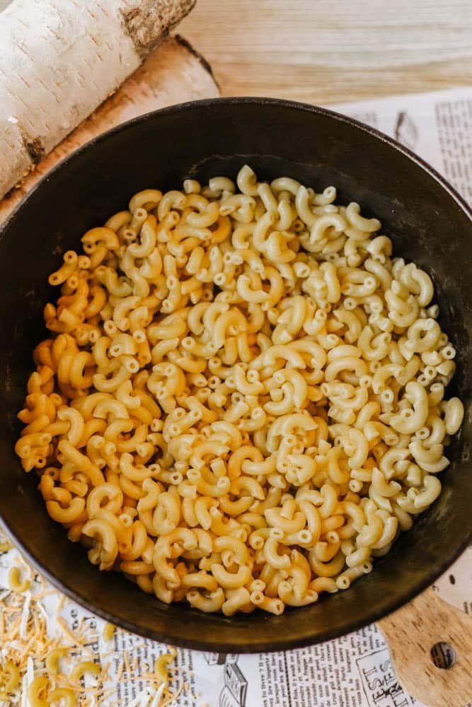 A black pot filled with cooked elbow macaroni sits on a table covered with a printed paper and some grated cheese scattered nearby.