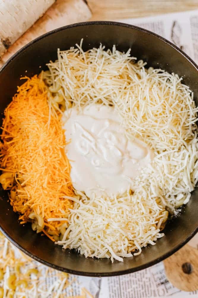 A pot filled with uncooked macaroni, topped with shredded cheddar and mozzarella cheese, and a dollop of creamy white sauce, ready to be mixed together.