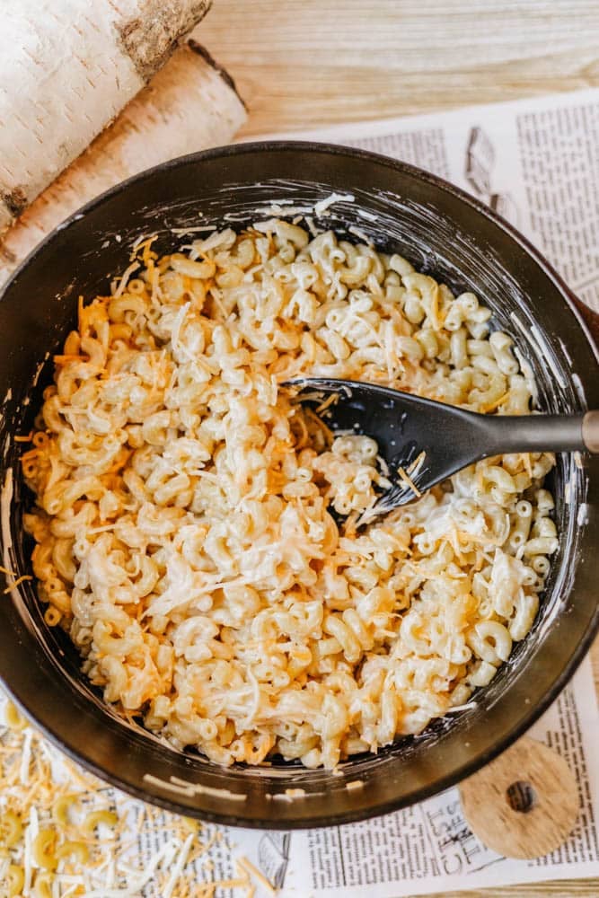 A black pot filled with creamy macaroni and cheese, being stirred with a black spoon. Grated cheese is visible on top, and the pot is placed on a table with newspaper underneath.