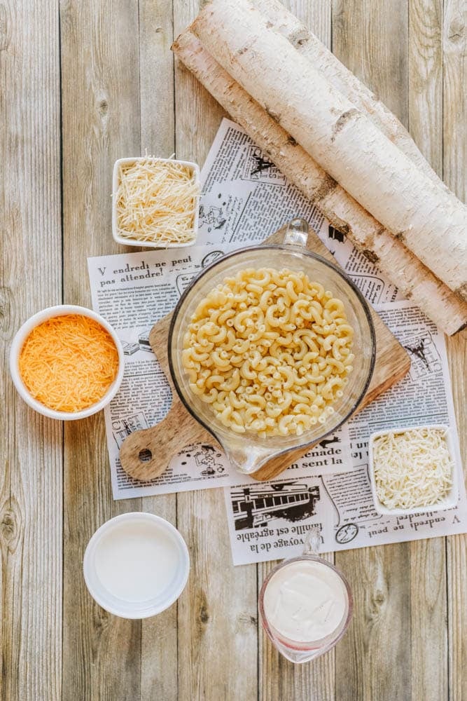 A glass bowl of uncooked macaroni sits on a wooden board with shredded cheeses, a cup of milk, and cream nearby, all placed on newspaper sheets over a wood table.
