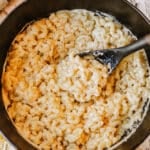 A black pot filled with creamy macaroni and cheese sits on a wooden surface, with a black serving spoon stirring the pasta. Shredded cheese is visible nearby.