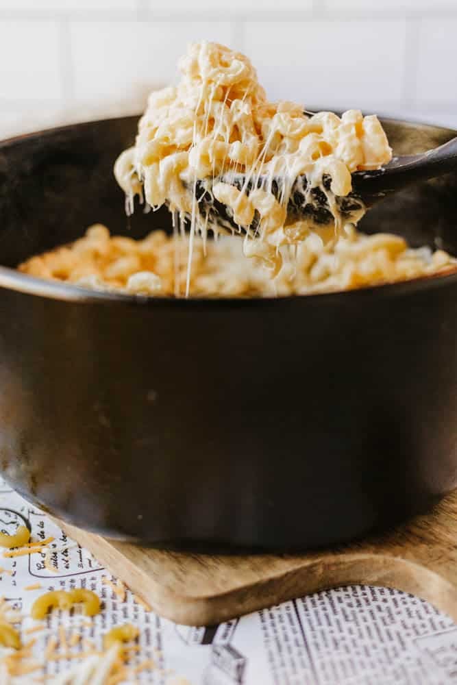 A spoon lifts a serving of cheesy macaroni and cheese from a black pot, with gooey melted cheese stretching from the dish. The pot sits on a wooden board atop a paper-covered surface.