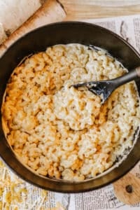 A black pot filled with creamy macaroni and cheese sits on a wooden surface, with a black serving spoon stirring the pasta. Shredded cheese is visible nearby.