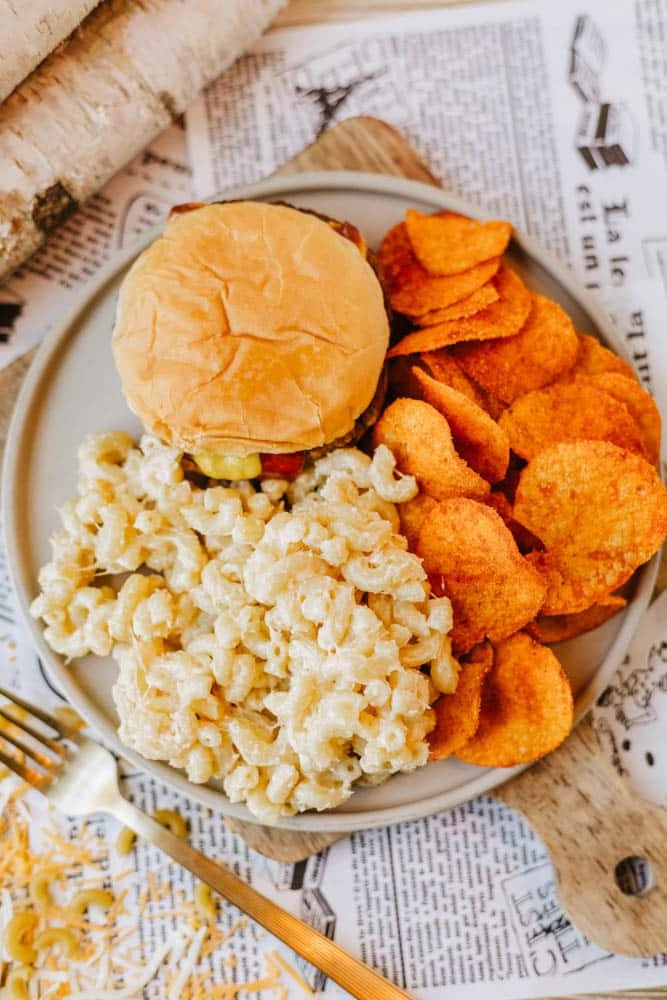 A plate with a cheeseburger, a serving of macaroni and cheese, and a pile of orange-colored potato chips sits on a table covered with printed paper. A gold fork and shredded cheese are nearby.