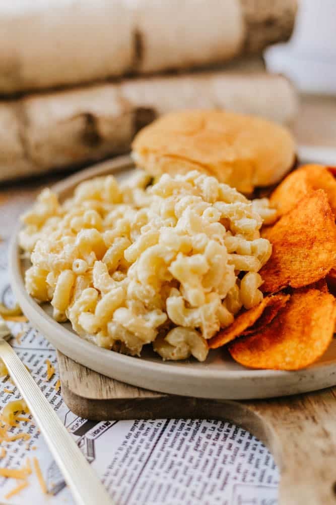 A plate of creamy macaroni and cheese, orange potato chips, and a sandwich bun sits on a wooden board with a fork, atop newsprint paper. Rolled napkins or logs are in the background.