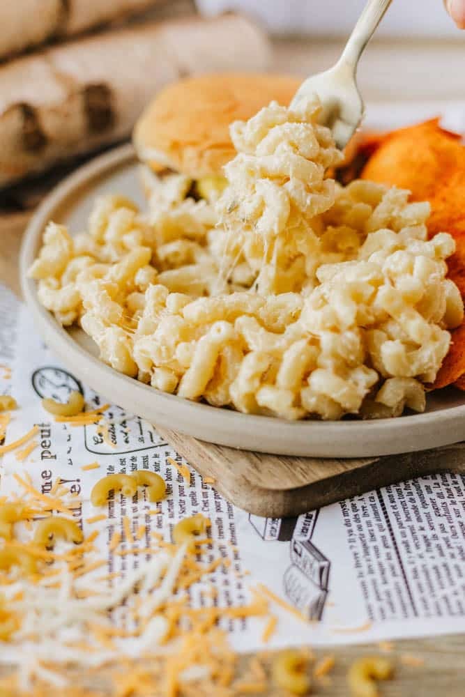 A fork lifts a cheesy scoop of macaroni and cheese from a plate, which also has a sandwich and orange chips. Shredded cheese and pasta are scattered on a newspaper underneath the plate.