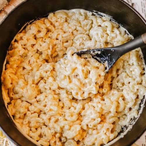 A black pot filled with creamy macaroni and cheese sits on a wooden surface, with a black serving spoon stirring the pasta. Shredded cheese is visible nearby.