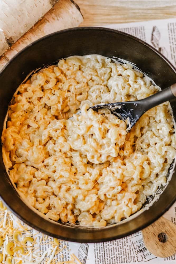 A black pot filled with creamy macaroni and cheese sits on a wooden surface, with a black serving spoon stirring the pasta. Shredded cheese is visible nearby.