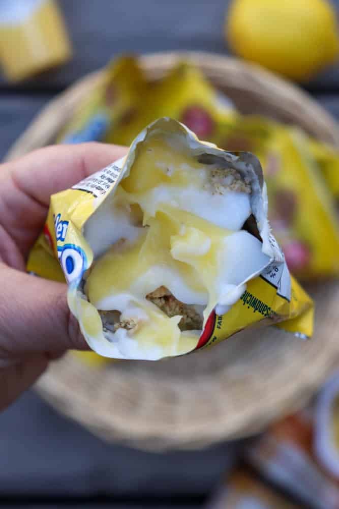 A hand holding an open granola bar wrapper filled with creamy yogurt and lemon curd, with a basket containing more granola bars blurred in the background.