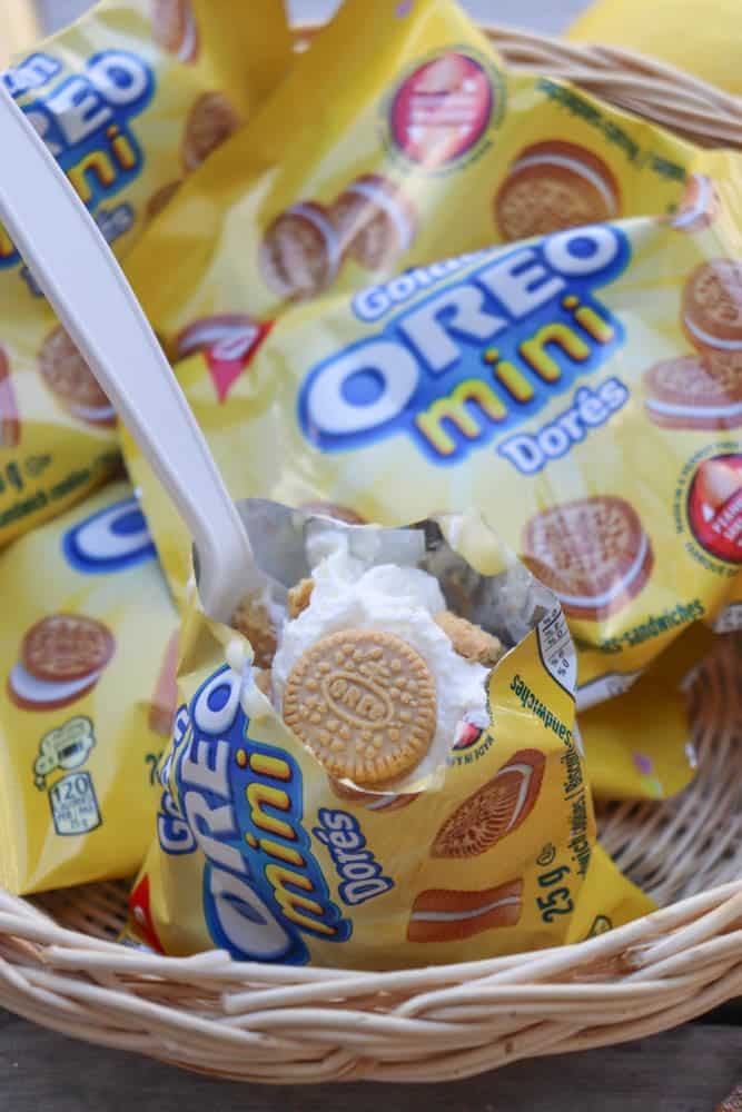 A plastic spoon scoops whipped cream and a golden mini Oreo from an open yellow packet of Oreo Mini Dorés cookies, with more unopened packets in a wicker basket underneath.