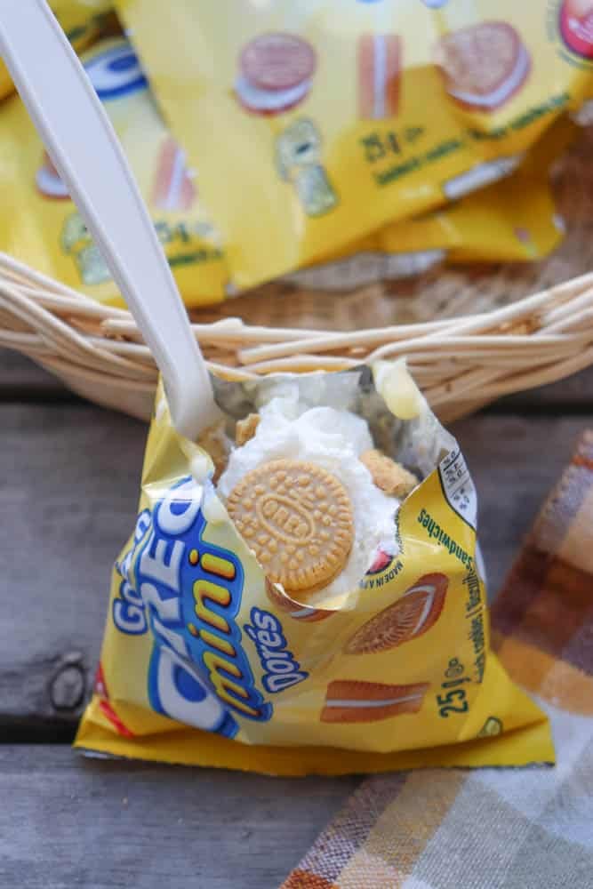 A small bag of Golden Oreo Mini cookies is open and filled with whipped cream and crumbled cookies, with a white plastic spoon inside. The bag rests on a wooden surface near a woven basket and a plaid cloth.