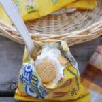 A small yellow packet of mini golden sandwich cookies is opened and filled with whipped cream, with a spoon sticking out. The packet sits in front of a wicker basket on a wooden surface next to a plaid cloth.