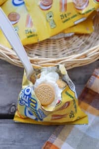 A small yellow packet of mini golden sandwich cookies is opened and filled with whipped cream, with a spoon sticking out. The packet sits in front of a wicker basket on a wooden surface next to a plaid cloth.