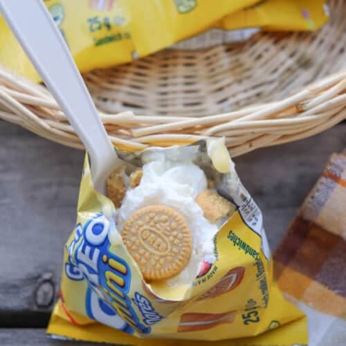 A small yellow packet of mini golden sandwich cookies is opened and filled with whipped cream, with a spoon sticking out. The packet sits in front of a wicker basket on a wooden surface next to a plaid cloth.