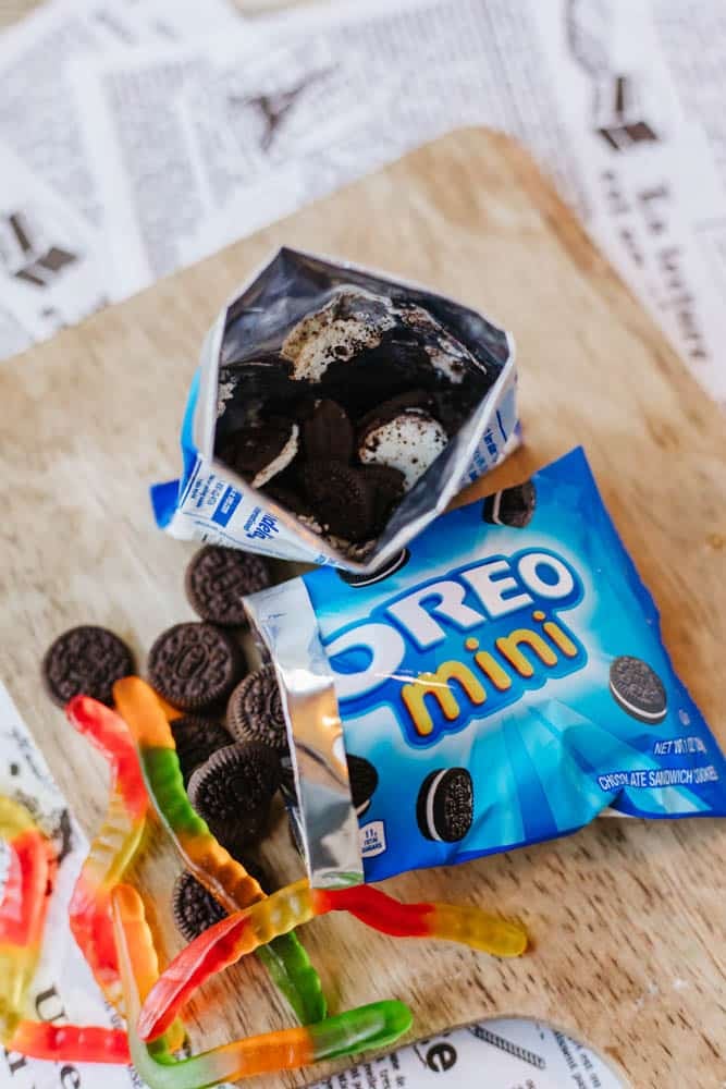 A blue package of Oreo Mini cookies is open on a wooden surface with some cookies spilling out. Colorful gummy worms are scattered nearby. There are pieces of newspaper in the background.