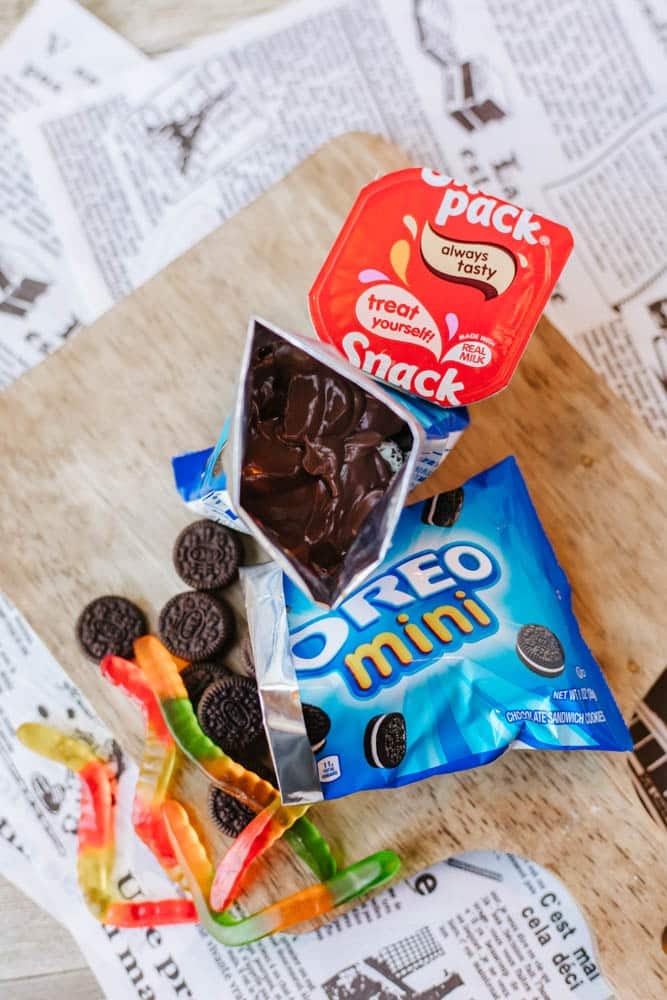 A cup of chocolate pudding, an open pack of Oreo minis, and colorful gummy worms are arranged on a wooden surface with torn newspaper pieces underneath.