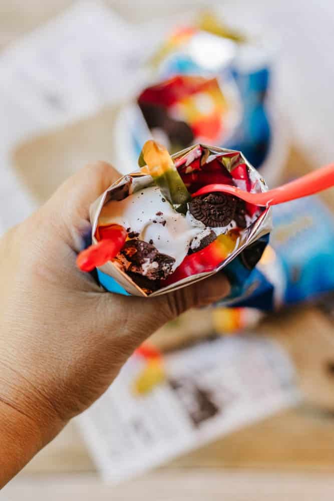 A hand holds an open chip bag filled with ice cream, crushed cookies, and colorful gummy worms, with a red spoon inside. Another similar treat is blurred in the background.