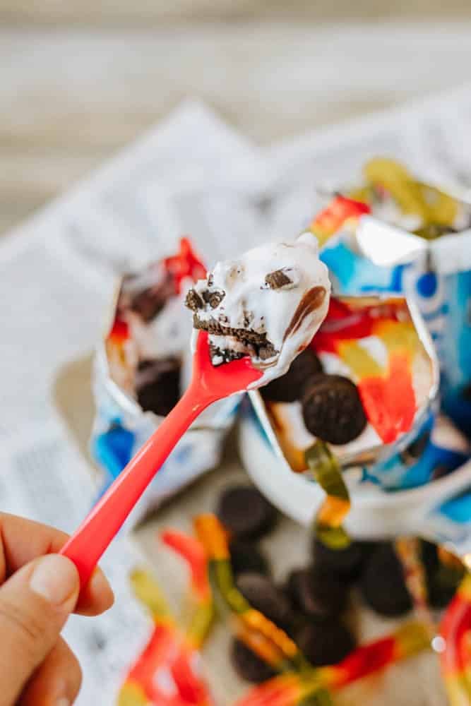 A hand holds a pink spoon with a bite of ice cream, crushed cookies, and chocolate from an open Oreo wrapper; gummy worms and mini cookies are scattered nearby.