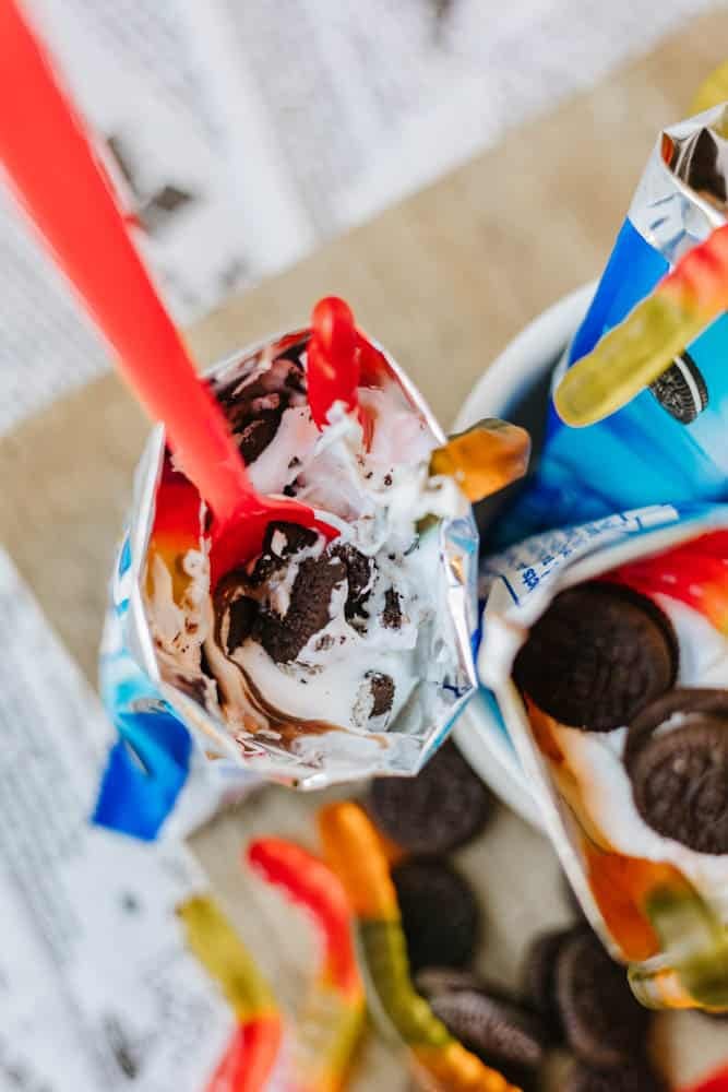 An open pudding cup topped with crushed chocolate cookies and colorful gummy worms, with a red spoon inside, sits on a surface scattered with more cookies and gummy worms.