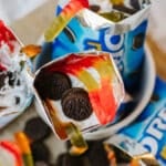 A close-up of an opened Oreo snack bag filled with whipped cream, mini Oreos, and colorful gummy worms, resembling a fun dessert parfait. Other similar bags and gummy worms are visible around it.