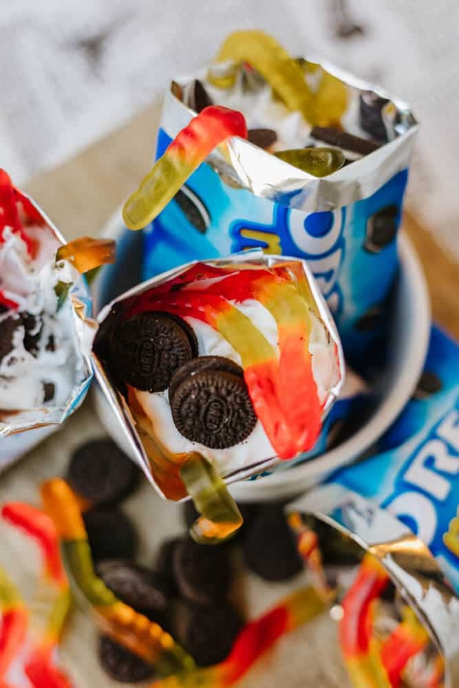 A close-up of an opened Oreo snack bag filled with whipped cream, mini Oreos, and colorful gummy worms, resembling a fun dessert parfait. Other similar bags and gummy worms are visible around it.